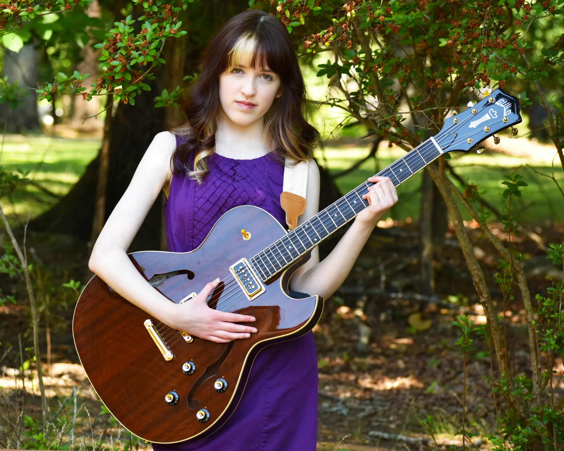 Josette Seraphine in a purple dress holding a hollow-body electric guitar among trees